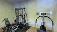 This single-floor fitness space is equipped to support a range of exercise needs within a straightforward, functional layout. The room features a multi-station cable strength machine prominently positioned against a textured, light-toned wall, complete with instructional exercise guides mounted directly on the unit for user reference. Additional posted guidance on the wall nearby further supports proper equipment use. A stair-climbing cardio machine with a digital display console is positioned to the right, offering cardiovascular training options alongside the strength equipment. The flooring transitions from a rubber mat beneath the strength equipment — providing stability and floor protection — to a carpet surface throughout the remainder of the room. The walls feature a vertically ribbed texture in neutral tones, contributing to a clean and understated aesthetic. The space is modest in scale but thoughtfully arranged to accommodate both strength and cardio training, making it a practical fitness facility suited for everyday exercise routines.