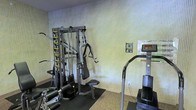 This single-floor fitness space is equipped to support a range of exercise needs within a straightforward, functional layout. The room features a multi-station cable strength machine prominently positioned against a textured, light-toned wall, complete with instructional exercise guides mounted directly on the unit for user reference. Additional posted guidance on the wall nearby further supports proper equipment use. A stair-climbing cardio machine with a digital display console is positioned to the right, offering cardiovascular training options alongside the strength equipment. The flooring transitions from a rubber mat beneath the strength equipment — providing stability and floor protection — to a carpet surface throughout the remainder of the room. The walls feature a vertically ribbed texture in neutral tones, contributing to a clean and understated aesthetic. The space is modest in scale but thoughtfully arranged to accommodate both strength and cardio training, making it a practical fitness facility suited for everyday exercise routines.