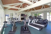 Located at 545 W Hacienda Ave in Campbell, California, this well-appointed fitness center offers a welcoming and functional environment for exercise and relaxation. The space is housed within a single-floor structure distinguished by striking vaulted ceilings with exposed wood beams and white structural framing, lending the interior a bright, airy, and architecturally appealing character. Track lighting runs along the ceiling, ensuring the space is well-lit throughout.

The main workout area features durable teal rubber flooring and is equipped with a comprehensive selection of cardio machines, including Cybex and Precor treadmills, ellipticals, and stationary bikes, as well as strength training equipment such as a Cybex multi-station machine and free weights with a wall-mounted mirror. Large windows and glass doors on multiple walls flood the space with natural light and provide views of surrounding trees and an outdoor pool area, creating an inviting atmosphere.

Adjacent to the workout zone is a comfortable lounge area featuring plush seating, a wall-mounted flat-screen television, a classic fireplace, and ceiling fans — offering a space to relax before or after a workout. The seamless flow between the lounge and gym areas, combined with the warm wood ceiling tones and abundant natural light, gives this fitness center a distinctive and welcoming character.