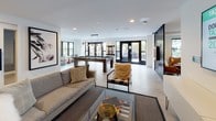The Club Suite is a thoughtfully designed amenity space that seamlessly blends entertainment, relaxation, and social gathering into one expansive single-floor layout. The open-concept design is anchored by a bright, airy lounge area featuring contemporary furnishings, including a plush sofa, accent chairs, and a wall-mounted flat-screen TV, all set against a clean neutral palette with polished concrete flooring. Black-framed glass partitions add a modern industrial touch while maintaining visual connectivity throughout the space. The game room is a standout feature, housing a full-size billiards table beneath pendant lighting, along with additional games such as shuffleboard and oversized Connect Four. Large black-framed windows and glass doors flood the space with natural light and offer views to the outdoors. A cozy dining nook is tucked within a richly colored teal-accented alcove, complete with a built-in banquette upholstered in deep navy, a round dining table, and mid-century modern chairs. Adjacent to the nook, a well-appointed kitchenette features warm wood cabinetry, subway tile backsplash, open shelving, stainless steel appliances, and a prep sink — ideal for hosting gatherings. Curated artwork throughout adds personality, while the cohesive design creates an inviting, club-like atmosphere.
