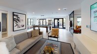 The Club Suite is a thoughtfully designed amenity space that seamlessly blends entertainment, relaxation, and social gathering into one expansive single-floor layout. The open-concept design is anchored by a bright, airy lounge area featuring contemporary furnishings, including a plush sofa, accent chairs, and a wall-mounted flat-screen TV, all set against a clean neutral palette with polished concrete flooring. Black-framed glass partitions add a modern industrial touch while maintaining visual connectivity throughout the space. The game room is a standout feature, housing a full-size billiards table beneath pendant lighting, along with additional games such as shuffleboard and oversized Connect Four. Large black-framed windows and glass doors flood the space with natural light and offer views to the outdoors. A cozy dining nook is tucked within a richly colored teal-accented alcove, complete with a built-in banquette upholstered in deep navy, a round dining table, and mid-century modern chairs. Adjacent to the nook, a well-appointed kitchenette features warm wood cabinetry, subway tile backsplash, open shelving, stainless steel appliances, and a prep sink — ideal for hosting gatherings. Curated artwork throughout adds personality, while the cohesive design creates an inviting, club-like atmosphere.