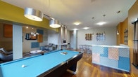 This single-floor amenity lounge is designed with a contemporary aesthetic, combining functionality and style in an open, well-lit environment. The centerpiece of the space is a full-sized pool table with a bright blue felt surface, positioned beneath a set of bold, industrial-style pendant lights that draw the eye and anchor the room's layout.

Warm hardwood flooring runs throughout, providing a rich contrast to the cool gray walls and modern furnishings. A comfortable lounge area is situated to one side, featuring a sofa and accent chair arranged around a low coffee table, ideal for casual conversation or relaxation. A computer workstation with a monitor and seating offers a practical workspace within the same open area.

On the opposite side, a bar-height counter with a textured decorative front and transparent acrylic stools serves as a social gathering spot. Colorful abstract wall art adds personality to the neutral palette. Pendant lighting and flush-mount ceiling fixtures provide even illumination throughout. The layout is open and thoughtfully organized, creating a versatile communal space suited for both entertainment and everyday use.