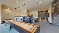 This single-floor amenity lounge is a well-appointed communal space designed for both recreation and relaxation. The open layout is anchored by a full-length shuffleboard table, making it an immediate focal point for social gatherings. Adjacent to the game area is a sleek wet bar finished in light cabinetry with dark countertops, featuring a stainless steel sink, a built-in beverage refrigerator branded "The Bond," and a microwave — fully equipped for entertaining. Overhead, a dramatic track lighting rail adds an industrial-modern touch to the bright, white ceiling. A casual dining nook with built-in banquette seating, round tables, and black metal chairs sits near large windows that invite natural light into the space. The lounge area is defined by a bold plaid accent wall that provides a striking backdrop for a generous sectional sofa in warm tan leather, complemented by teal and gray throw pillows and a large wall-mounted flat-screen television. Carpet tile flooring in a contemporary pattern runs throughout. A corridor with dramatic dark stone tile cladding leads to a frosted-glass private meeting room, adding a professional element to this multifunctional space. The overall ambiance is modern, vibrant, and inviting.