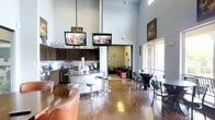 This single-floor communal space is designed for both socializing and relaxation, offering a well-appointed layout that combines a kitchenette, bar-height seating, and multiple lounge areas. The soaring double-height ceilings immediately establish a sense of openness, while two flat-screen televisions suspended from the ceiling on drop mounts serve as a central entertainment focal point, visible from nearly every angle of the room.

The kitchenette area features dark espresso cabinetry, a microwave, coffee station, and a curved bar counter complemented by modern adjustable barstools. The polished concrete floors reflect ambient light throughout, adding an industrial-modern aesthetic that ties the space together cohesively.

Dining-height tables with black chairs are positioned near a window that introduces natural light, creating a bright and functional area for casual meals or work. In the background, a connected lounge room is visible, furnished with sofas and accent chairs for quieter, more relaxed gatherings. Decorative wall art and subtle pendant lighting complete the space, lending it a curated, contemporary feel suited for everyday community use.