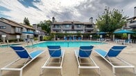 Located in Roseville, California, this community pool area at 3 Somer Ridge Dr offers a well-appointed outdoor retreat designed for relaxation and recreation. The centerpiece of the space is a generously sized swimming pool with clear, blue water, complemented by a designated lap lane for fitness-minded swimmers. The pool deck is finished in a textured aggregate surface, providing both durability and slip resistance, and is outfitted with multiple blue-and-white lounge chairs positioned to take full advantage of the California sun.

The surrounding area includes shaded seating zones with blue patio umbrellas and tables, ideal for casual gatherings. A secondary, freeform pool is also present on the property, offering a more leisurely swim experience. The pool complex is secured by a black iron gate and fence, ensuring a safe and private environment for residents.

Adjacent to the pool is a community building featuring a distinctive circular window detail and covered colonnade, which appears to house support facilities for the pool area. Mature trees, including tall pines and deciduous varieties, frame the space and provide natural shade and privacy. The multi-story residential buildings visible in the background feature balconies and chimneys, contributing to the cohesive architectural character of the community. The overall ambiance is clean, inviting, and well-maintained.