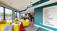 Located at 2144 E Republic Rd in Springfield, Missouri, this contemporary single-floor space is designed with a bold, modern aesthetic and a strong sense of community in mind. The open-plan layout seamlessly connects the lounge, dining, and kitchen areas, creating a cohesive and welcoming environment throughout.

The lounge area makes an immediate impression with vibrant yellow armchairs, a colorful area rug, and floor-to-ceiling corner windows that flood the space with natural light and offer sweeping views of the surrounding greenery and outdoor terrace. The teal accent walls add a striking visual element that carries through the space, lending it a dynamic and energetic character.

The fully equipped kitchen features stainless steel appliances, including a full-size refrigerator, stove, and microwave, complemented by two-tone cabinetry in light wood and dark wood-grain finishes. A large dark-countertop island with pendant lighting above serves as both a prep area and casual gathering spot, with additional seating along its length.

Adjacent to the kitchen, the dining area is anchored by a rustic wooden table with modern black chairs, positioned beside a large window that opens to a balcony overlooking mature trees. The combination of contemporary finishes, bold color choices, abundant natural light, and direct outdoor access makes this a highly functional and visually engaging shared space.