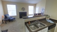 This single-floor residence offers a comfortable and functional open-concept design that seamlessly connects the kitchen and living areas. The kitchen is equipped with dark granite-style countertops, white cabinetry, and a double stainless steel sink, providing ample workspace for everyday use. A peninsula counter extends into the living space, enhancing the open flow between the two areas and offering additional prep or serving space.

The living room is carpeted and bathed in natural light, thanks to multiple large windows that line the walls and allow sunlight to fill the space throughout the day. A classic fireplace with dark tile surround and a clean white mantle serves as the room's focal point, adding warmth and character to the overall ambiance. The neutral, warm-toned walls throughout create a cohesive and inviting atmosphere.

The layout is well-suited for both everyday living and casual entertaining, with the kitchen and living room maintaining a strong visual connection. The single-floor plan ensures easy navigation throughout the home, making it practical and accessible. Overall, the space combines functional design with comfortable living, highlighted by abundant natural light, a traditional fireplace, and thoughtfully arranged living areas.