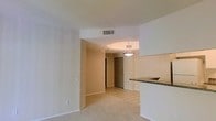 This single-floor residence offers a thoughtfully arranged open layout that seamlessly connects the main living spaces. The living area features soft wall-to-wall carpeting, creating a warm and comfortable atmosphere, while the entryway and adjoining spaces transition to light-toned ceramic tile flooring for easy maintenance.

A defining feature of the layout is the arched passageway that frames the transition between the living area and the tiled entryway corridor, adding a subtle architectural detail to the otherwise clean, neutral interior. A pendant light fixture hangs above the entryway, providing a focal point and functional illumination to the space.

The kitchen is positioned adjacent to the living area and is distinguished by a raised breakfast bar topped with granite-style countertops, offering a natural division between the kitchen and living space while maintaining an open, connected feel. The white cabinetry contributes to a bright and airy aesthetic throughout.

Smooth, light-colored walls and white trim run throughout the unit, complemented by a ceiling-mounted HVAC vent that ensures year-round climate control. The overall ambiance is clean, understated, and move-in ready, making it a practical and comfortable living environment.