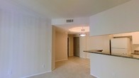 This single-floor residence offers a thoughtfully arranged open layout that seamlessly connects the main living spaces. The living area features soft wall-to-wall carpeting, creating a warm and comfortable atmosphere, while the entryway and adjoining spaces transition to light-toned ceramic tile flooring for easy maintenance.

A defining feature of the layout is the arched passageway that frames the transition between the living area and the tiled entryway corridor, adding a subtle architectural detail to the otherwise clean, neutral interior. A pendant light fixture hangs above the entryway, providing a focal point and functional illumination to the space.

The kitchen is positioned adjacent to the living area and is distinguished by a raised breakfast bar topped with granite-style countertops, offering a natural division between the kitchen and living space while maintaining an open, connected feel. The white cabinetry contributes to a bright and airy aesthetic throughout.

Smooth, light-colored walls and white trim run throughout the unit, complemented by a ceiling-mounted HVAC vent that ensures year-round climate control. The overall ambiance is clean, understated, and move-in ready, making it a practical and comfortable living environment.