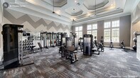 This fitness center in Durham, NC offers a generously sized, single-floor layout designed to accommodate a full range of workout activities. The space is defined by its striking architectural details, including an elegant coffered ceiling with recessed lighting and pendant fixtures that lend the room a refined, upscale character uncommon in typical gym settings. A bold gray-and-white chevron pattern runs along the upper walls, adding a dynamic visual element that energizes the environment.

Large, multi-pane windows line the perimeter walls, flooding the interior with natural light and providing views of the surrounding outdoor landscape. The openness created by the tall ceilings and abundant glazing contributes to an airy, motivating atmosphere. Textured, patterned carpet tile in muted gray and blue tones covers the floor throughout, providing both durability and a modern aesthetic.

The main workout area is equipped with a comprehensive selection of strength training machines, including cable systems, selectorized weight machines, and an Octagon functional trainer, as well as adjustable benches and a full dumbbell rack backed by floor-to-ceiling mirrors. A dedicated cardio zone features treadmills, ellipticals, and stair climbers positioned in front of the expansive windows. An additional room accommodates cycling bikes, stability balls, and storage. Ceiling fans and mounted TVs enhance comfort and usability throughout.