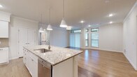 This well-appointed single-floor residence offers a thoughtfully designed open-concept layout that seamlessly connects the kitchen, dining, and living areas. The kitchen serves as the heart of the home, anchored by a generous granite-topped island with a built-in sink and pendant lighting overhead. White shaker-style cabinetry lines the walls, complemented by a classic subway tile backsplash, stainless steel appliances including a refrigerator, microwave, and range, and recessed lighting throughout. Warm hardwood-style flooring extends across the main living areas, enhancing the bright and airy ambiance. The living area is bathed in natural light from large sliding glass doors and dual windows, with crown molding adding an elegant architectural touch. The bedroom is spacious and carpeted, featuring multiple windows with abundant natural light and a ceiling fan. The bathrooms are finished to a high standard — one features a granite vanity and tiled tub surround with a glass block window, while the other showcases a walk-in glass-enclosed shower with marble-style subway tile walls, penny tile flooring, and a granite vanity. Together, these spaces create a cohesive, refined living environment with quality finishes at every turn.