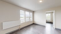 This efficiently designed 3-bedroom, 1-bathroom unit offers a clean, modern aesthetic across a single floor. The interior features durable grey laminate flooring and neutral cream-toned walls throughout, creating a cohesive and easy-to-maintain living environment. Each bedroom is equipped with panel radiators for heating and fitted with horizontal-slatted window blinds that allow natural light while maintaining privacy. The primary bedroom includes a built-in closet with dual hanging rods, offering organized storage. One bedroom features a glazed exterior door providing direct outdoor access. The living room is bright and open, with multiple windows overlooking the surrounding area. The kitchen is outfitted with light wood-grain cabinetry, light stone-finish countertops, a stainless steel sink, a dishwasher, and a full-size refrigerator — providing functional efficiency in a compact footprint. The bathroom features a pedestal sink, toilet, and wood-tone flooring, maintaining the unit's clean, minimal style. A dedicated laundry room houses a full-size Samsung front-loading washer and dryer set. Overall, the unit presents a practical, well-appointed layout suited for multi-occupant living.