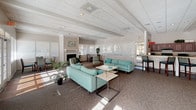 This single-floor community clubhouse offers a generously proportioned and well-appointed gathering space designed for both relaxation and functionality. The heart of the space is a expansive open lounge area with high beamed ceilings, a textured tile ceiling, and abundant natural light streaming through large windows and multiple sets of French doors that look out onto a pool area. The lounge is furnished with teal upholstered sofas and accent chairs arranged around low coffee tables, creating comfortable conversation areas. A classic fireplace with a decorative surround serves as a focal point along one wall, flanked by additional seating and dining arrangements, adding warmth and character to the room. The carpeted floors throughout the main area contribute to a comfortable, welcoming ambiance. A dedicated workstation area with computers and office equipment occupies one corner of the lounge, providing a practical workspace for administrative or communal use. Adjacent to the main lounge is a functional kitchen, featuring rich dark wood cabinetry, light wood-look flooring, black appliances including a full-size refrigerator and gas range, and generous countertop workspace. The kitchen is well-equipped for catering and community events. Tropical potted plants placed throughout the space add a fresh, inviting touch to the overall atmosphere.
