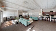 This single-floor community clubhouse offers a generously proportioned and well-appointed gathering space designed for both relaxation and functionality. The heart of the space is a expansive open lounge area with high beamed ceilings, a textured tile ceiling, and abundant natural light streaming through large windows and multiple sets of French doors that look out onto a pool area. The lounge is furnished with teal upholstered sofas and accent chairs arranged around low coffee tables, creating comfortable conversation areas. A classic fireplace with a decorative surround serves as a focal point along one wall, flanked by additional seating and dining arrangements, adding warmth and character to the room. The carpeted floors throughout the main area contribute to a comfortable, welcoming ambiance. A dedicated workstation area with computers and office equipment occupies one corner of the lounge, providing a practical workspace for administrative or communal use. Adjacent to the main lounge is a functional kitchen, featuring rich dark wood cabinetry, light wood-look flooring, black appliances including a full-size refrigerator and gas range, and generous countertop workspace. The kitchen is well-equipped for catering and community events. Tropical potted plants placed throughout the space add a fresh, inviting touch to the overall atmosphere.
