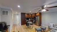 This single-floor residence offers a thoughtfully designed open-concept layout that seamlessly blends living and dining areas into one cohesive space. The kitchen is a standout feature, showcasing rich, dark-toned wood cabinetry that extends floor to ceiling for generous storage, paired with sleek black countertops and a full suite of stainless steel appliances, including a French door refrigerator and a professional-style gas range. Recessed lighting is installed throughout the ceiling, providing bright, even illumination across the space. A ceiling fan adds both functionality and a subtle decorative touch. The dining area sits adjacent to the kitchen and is furnished with a contemporary glass-top table and modern upholstered chairs, making it an ideal spot for everyday meals or entertaining. Warm-toned hardwood flooring runs continuously throughout the space, contributing to a cohesive and inviting ambiance. A hallway extending from the main living area leads to additional rooms, each accessible through classic white paneled doors. The overall design balances practicality with a clean, modern aesthetic, creating a comfortable and functional living environment.