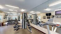 Located at 334 Carolina Forest Blvd in Jacksonville, North Carolina, this fitness center offers a comprehensive workout facility designed to accommodate a wide range of exercise needs. The single-floor space features durable speckled rubber flooring throughout, providing both safety and comfort during workouts. Large wall-to-wall mirrors line multiple walls, enhancing the sense of space and allowing users to monitor their form. Bright fluorescent lighting, complemented by ceiling fans, ensures the area remains well-lit and comfortable during use. Several windows allow natural light to filter in, contributing to an open and airy atmosphere. The cardio section is equipped with multiple treadmills, elliptical trainers, and stationary bikes, each outfitted with individual display monitors. The strength training area features a multi-station cable machine along with a well-stocked dumbbell rack offering a full range of free weights. Wall-mounted televisions provide entertainment throughout the space. Additional amenities include a water cooler station for hydration convenience and informational boards mounted near the entrance. The facility is equipped with clearly marked emergency exits and security cameras, prioritizing user safety. Overall, the space provides a functional and well-organized environment suited for both cardio and strength-focused fitness routines.