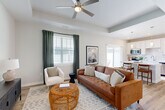 This thoughtfully designed one-bedroom, one-bathroom residence in Charlotte, NC offers an efficient and comfortable layout with modern finishes throughout. The entrance welcomes you with a decorative wall-mounted shelf and mirror, leading through a hallway into the main living areas. The open-concept living room features a tray ceiling with a ceiling fan, light-toned hardwood flooring, and large windows dressed with elegant dark green curtains that allow generous natural light to fill the space. The kitchen is a standout feature, showcasing white shaker cabinetry, stainless steel appliances, a center island with seating for two, and pendant lighting overhead. A sliding glass door in the kitchen provides access to an outdoor area. The bedroom offers a comfortable retreat with dual double-door closets, a bright window with natural light, and hardwood flooring. The bathroom is clean and functional, featuring a full soaking tub and shower combination, a vanity with white cabinetry, and a window for natural light. Consistent light wood flooring and a neutral color palette tie the entire residence together, creating a cohesive, modern aesthetic throughout.