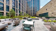 This expansive outdoor courtyard terrace is nestled within a contemporary urban development, enclosed on multiple sides by striking multi-story buildings clad in textured brick and floor-to-ceiling glazing. The terrace is thoughtfully organized into several distinct zones, each catering to a different style of outdoor use.

A generous turfed lawn area serves as the centerpiece, providing an open, flexible space accented by colorful modular seating poufs and freestanding planter bowls filled with lush greenery. Adjacent to the lawn, a hardscaped patio zone features dining tables and teal chairs suited for al fresco meals, alongside a dedicated lounge area furnished with woven rope armchairs and plush sofas layered with vibrant cushions, arranged atop decorative outdoor rugs.

A timber-decked walkway runs along one side of the terrace, bordered by long, steel-framed planters brimming with flowering and leafy plants set against the building's warm brick façade and large steel-framed windows. Raised planting beds throughout the space introduce trees, shrubs, and seasonal greenery, creating a sense of natural enclosure within the urban setting.

The courtyard benefits from open-sky natural light, and the surrounding architecture provides a dramatic vertical backdrop that defines the space. Wall-mounted lighting and overhead string-light rigging suggest usability into the evening hours.