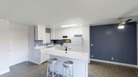 This single-floor space centers around a well-appointed kitchen designed with both functionality and style in mind. The kitchen showcases crisp white shaker-style cabinetry that extends to the upper walls, offering generous storage throughout. A flush-mounted ceiling light brightens the cooking area, complementing the natural brightness of the space. White appliances, including a refrigerator, electric range, microwave, and dishwasher, are seamlessly integrated into the layout for a cohesive look. A double-basin sink with a modern faucet is positioned along the perimeter counter, adding practicality to the workspace. The kitchen's peninsula island serves as a focal point, featuring an expansive white countertop that provides additional prep space and casual dining with two sleek metallic bar stools. The color scheme pairs clean white surfaces with bold slate-blue accent walls, creating a modern, contrast-driven aesthetic. Dark wood-look flooring grounds the space and adds warmth, balancing the otherwise cool tones throughout. The open layout between the kitchen and adjoining area contributes to a spacious, airy ambiance well-suited for everyday living and entertaining.