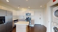 This contemporary 2-bedroom, 2-bathroom residence at 10 DeKalb Avenue in White Plains, NY offers a thoughtfully designed open-concept layout with modern finishes throughout. Rich dark hardwood flooring flows seamlessly across the entire living space, complementing the clean white walls and recessed lighting overhead.

The kitchen is a standout feature, boasting sleek white upper cabinetry, warm wood-toned lower cabinets, and bright white countertops. A full suite of stainless steel appliances — including a refrigerator with ice maker, range, microwave, and dishwasher — is anchored by a freestanding center island, ideal for additional prep space or casual dining.

The living area is bathed in natural light courtesy of expansive floor-to-ceiling windows with sliding glass doors that open to a balcony, offering open views of the surrounding White Plains neighborhood. Both bedrooms are generously proportioned, featuring the same hardwood flooring, large windows with outdoor views, and ample closet space.

Each of the two bathrooms is finished with large-format tile flooring, white subway tile surrounds, and a decorative mosaic accent band above the soaking tub, providing a clean and polished aesthetic. Recessed lighting throughout enhances the bright, modern ambiance of this well-appointed residence.