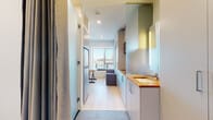 This thoughtfully designed single-floor unit maximizes every inch of its compact footprint through smart layout planning and modern finishes. The space flows from an entry corridor into a combined living and kitchen area, where light tile flooring transitions to warm wood planks underfoot. The kitchenette is a standout feature, showcasing a rich butcher block countertop that contrasts beautifully with sleek white cabinetry. A stainless steel undermount sink with a pull-down faucet and an integrated cooktop with overhead exhaust hood complete the functional cooking area. A mirrored wall cabinet adds brightness and utility near the sink zone. The living area is anchored by a large window that frames an urban cityscape, flooding the space with natural light and providing an airy, open feel. Built-in cabinetry along one wall offers generous storage solutions, keeping the living space uncluttered. A wall-mounted air conditioning unit ensures year-round climate comfort. The bathroom is cleanly appointed with tile flooring and straightforward fixtures. Recessed ceiling lighting throughout maintains a modern, polished aesthetic. The overall design is minimal yet practical, making excellent use of the available space.