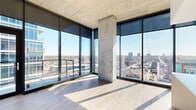 Located at 81 E 14th St in the heart of Kansas City, MO, this high-rise unit showcases a contemporary design defined by soaring floor-to-ceiling windows that frame striking panoramic views of the downtown skyline and surrounding cityscape. Exposed concrete columns and ceilings lend an industrial-modern character, while light-toned hardwood flooring runs throughout the open living areas, creating a warm contrast to the raw structural elements.

The kitchen is a standout feature, centered around a large island with a crisp white quartz countertop and an integrated sink. Cabinetry combines high-gloss white and natural wood-toned finishes, complemented by a glass tile backsplash, stainless steel appliances including a refrigerator and range, and a built-in microwave. Track lighting and a statement drum pendant illuminate the space effectively.

The living areas benefit enormously from corner-wrapped glazing, flooding the interior with natural light and offering unobstructed city views. Bedroom spaces continue the floor-to-ceiling window theme, with exposed ductwork adding to the industrial aesthetic.

Bathrooms feature marble-style tile throughout, including a glass-enclosed shower and a separate soaking tub, paired with a modern floating vanity. The overall ambiance is clean, bright, and sophisticated.