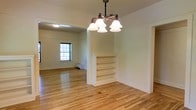This well-appointed two-floor space combines classic architectural charm with functional design. The main living area immediately draws attention with its warm, light-toned hardwood floors that extend throughout, lending a sense of continuity and warmth to the space. A decorative chandelier with bell-shaped shades hangs as a focal point, adding a touch of timeless elegance to the room.

A built-in shelving unit serves as a natural room divider, offering both practical storage and a distinctive architectural feature. The open layout allows for generous, flexible use of the living and dining areas, with ample room for a variety of furnishing arrangements.

Natural light filters in through a classic multi-pane window, brightening the interior and complementing the soft, neutral wall tones. A baseboard heating unit is discreetly positioned along the wall, ensuring year-round comfort. The clean white walls and trim throughout provide a crisp, versatile backdrop.

Spanning two floors, the space offers well-defined zones for living and private use, making it both practical and comfortable. The overall ambiance is one of understated classic character, with thoughtful built-in details and durable finishes that speak to quality and lasting style.