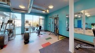 This fitness center offers a comprehensive and energizing workout environment designed to support a full range of exercise routines. The space is defined by its bold teal walls, which create a vibrant and motivating atmosphere, while the light-colored flooring is accented with colorful rectangular inlays in blue, green, orange, and red, adding a dynamic visual element throughout.

The cardio zone is well-stocked with multiple treadmills, an elliptical trainer, a stationary bike, and a rowing machine, providing ample options for aerobic workouts. A dedicated strength training area features a multi-function cable machine, a weight bench, and a full rack of dumbbells and kettlebells in graduated sizes, catering to users of all fitness levels. Large wall-mounted mirrors span one section of the space, enhancing the sense of openness and allowing users to monitor their form during workouts.

Exposed industrial-style ductwork runs across the ceiling, complementing the modern aesthetic, while flush-mounted circular light fixtures provide consistent, even illumination throughout. Large windows along one wall invite natural light and offer views to an adjacent outdoor area, creating an open and airy feel. The flooring transitions between smooth light panels and rubberized matting in the free weights zone for added safety and comfort. The space is accessible via both a single and double door entry.