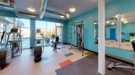 This fitness center offers a comprehensive and energizing workout environment designed to support a full range of exercise routines. The space is defined by its bold teal walls, which create a vibrant and motivating atmosphere, while the light-colored flooring is accented with colorful rectangular inlays in blue, green, orange, and red, adding a dynamic visual element throughout.

The cardio zone is well-stocked with multiple treadmills, an elliptical trainer, a stationary bike, and a rowing machine, providing ample options for aerobic workouts. A dedicated strength training area features a multi-function cable machine, a weight bench, and a full rack of dumbbells and kettlebells in graduated sizes, catering to users of all fitness levels. Large wall-mounted mirrors span one section of the space, enhancing the sense of openness and allowing users to monitor their form during workouts.

Exposed industrial-style ductwork runs across the ceiling, complementing the modern aesthetic, while flush-mounted circular light fixtures provide consistent, even illumination throughout. Large windows along one wall invite natural light and offer views to an adjacent outdoor area, creating an open and airy feel. The flooring transitions between smooth light panels and rubberized matting in the free weights zone for added safety and comfort. The space is accessible via both a single and double door entry.