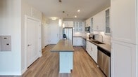 This single-floor residence offers a thoughtfully designed open layout with consistent wood-look flooring flowing throughout the main living areas. The kitchen serves as a central highlight, featuring white shaker-style cabinetry with glass-front upper cabinets, dark countertops, a subway tile backsplash, and a full suite of stainless steel appliances including a refrigerator with ice dispenser, electric range, over-the-range microwave, and dishwasher. A substantial center island with pendant lighting overhead provides additional counter space and casual seating. The living area is generously sized, benefiting from large, multi-pane windows that bring in abundant natural light and offer exterior views. A ceiling fan adds both functionality and comfort to the space. The bedroom is finished with plush wall-to-wall carpeting, a ceiling fan, and a window with exterior views, creating a comfortable retreat. The bathroom is well-appointed with wood-look flooring, a white vanity with dark countertops, and a tub-shower combination featuring large-format tile surround. Neutral tones and clean finishes are carried consistently throughout, contributing to a cohesive and modern aesthetic across the entire space.