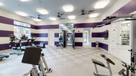 Located at 103 Inspiration Ave in Bluffton, South Carolina, this single-floor fitness center is designed to provide a comprehensive workout experience in a vibrant, well-appointed setting. The facility is immediately distinguished by its bold décor — wide horizontal stripes of deep purple and crisp white span the walls throughout, giving the space an energetic and motivating atmosphere.

The main workout area is generously proportioned and bathed in bright, even light from large recessed panel fixtures mounted across a white ceiling, complemented by multiple ceiling fans that promote consistent airflow. A row of treadmills and elliptical trainers lines one wall, each equipped with individual display screens, while full-length mirrors expand the sense of space and allow users to monitor their form. Strength training is well represented, with a multi-station cable machine, leg press, and various selectorized weight machines positioned throughout.

A separate functional training room offers a distinct environment, featuring lighter walls accented with playful polka-dot wall decor, natural light from multiple windows overlooking greenery, and a fully stocked functional training rig with resistance bands, kettlebells, a punching bag, and a rowing machine — ideal for dynamic and cross-training workouts.

Durable checkered tile flooring runs throughout the facility, and exterior-facing windows and glass doors bring in natural light and views of the surrounding landscape.