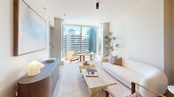 Located in the heart of Seattle's Belltown neighborhood, this thoughtfully designed one-bedroom, one-bathroom residence offers an efficient yet refined living experience across a single floor. The open-plan living, dining, and kitchen area serves as the centerpiece of the home, where light wood flooring, neutral tones, and clean lines create a cohesive modern aesthetic. Floor-to-ceiling windows in the living room flood the space with natural light while framing striking views of the Seattle skyline, making the area feel open and airy despite its compact footprint.

The kitchen is outfitted with flat-front cabinetry in a soft grey finish, an integrated cooktop, under-cabinet lighting, and a central island that doubles as additional prep space and casual dining. Built-in open shelving adds both storage and character. A dedicated dining area with pendant lighting sits adjacent to the kitchen, completing the social hub of the home.

The bedroom features large windows with city views, providing ample natural light, and includes closet space for practical storage. The bathroom is finished with elegant marble-style tile surrounding a soaking tub, accented by matte black fixtures for a contemporary touch. A dedicated laundry area adds everyday convenience to this polished urban residence.