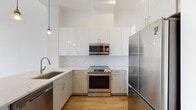 Located at 1400 Main St in Weymouth, MA, this thoughtfully designed single-floor residence combines modern finishes with a clean, open aesthetic. The kitchen is a standout feature, showcasing a two-tone cabinet design with high-gloss white upper cabinets and light wood-grain lower cabinets, complemented by a classic white subway tile backsplash. Stainless steel appliances — including a refrigerator, electric range, built-in microwave, and dishwasher — add a polished, professional quality. Pendant lighting and a undermount sink complete the well-appointed space. An open-concept layout connects the kitchen to the living area via a breakfast bar peninsula, creating a seamless flow ideal for entertaining. Warm hardwood-style flooring runs consistently throughout the unit, contributing to the cohesive, contemporary feel. The bedrooms are bright and airy, with tall black-framed windows that frame views of the surrounding landscape and invite generous natural light. Additional storage is provided by a dedicated closet space. The bathroom features large-format tile flooring, a tiled soaking tub and shower combination, a light wood vanity with a white quartz countertop, and elegant wall sconce lighting above a frameless mirror — offering a clean, spa-like atmosphere.
