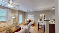 This single-floor residence is thoughtfully designed with a cohesive, neutral color palette of warm greige tones throughout, creating a calm and inviting atmosphere. The open-concept main living area seamlessly connects the living room, dining area, and kitchen, promoting an airy and functional flow. The living room features warm hardwood-style flooring, a ceiling fan, and large windows that allow generous natural light to fill the space. A comfortable seating arrangement anchors the room, complemented by a classic wooden coffee table.

The dining area sits centrally within the open layout and accommodates a round table with upholstered seating, illuminated by a sleek track lighting fixture overhead. Adjacent to the dining space, a breakfast bar with tall seating provides an additional dining or casual gathering option, connecting directly to the kitchen.

The galley-style kitchen is well-equipped with rich dark wood cabinetry, stone-toned countertops, black appliances including a range, microwave, and refrigerator, and a double-basin sink. Light flooring throughout the kitchen enhances brightness in the space.

The bedroom offers a cozy retreat with soft carpeting, warm neutral walls, and ample space for a full bedroom suite. The bathroom features dark wood vanity cabinetry, a large mirror, and stylish overhead vanity lighting, rounding out this comfortable and practical residence.