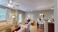 This single-floor residence is thoughtfully designed with a cohesive, neutral color palette of warm greige tones throughout, creating a calm and inviting atmosphere. The open-concept main living area seamlessly connects the living room, dining area, and kitchen, promoting an airy and functional flow. The living room features warm hardwood-style flooring, a ceiling fan, and large windows that allow generous natural light to fill the space. A comfortable seating arrangement anchors the room, complemented by a classic wooden coffee table.

The dining area sits centrally within the open layout and accommodates a round table with upholstered seating, illuminated by a sleek track lighting fixture overhead. Adjacent to the dining space, a breakfast bar with tall seating provides an additional dining or casual gathering option, connecting directly to the kitchen.

The galley-style kitchen is well-equipped with rich dark wood cabinetry, stone-toned countertops, black appliances including a range, microwave, and refrigerator, and a double-basin sink. Light flooring throughout the kitchen enhances brightness in the space.

The bedroom offers a cozy retreat with soft carpeting, warm neutral walls, and ample space for a full bedroom suite. The bathroom features dark wood vanity cabinetry, a large mirror, and stylish overhead vanity lighting, rounding out this comfortable and practical residence.