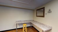 This single-floor space is designed with functionality and practicality at its core. The layout includes at least two distinct areas, each serving a specific purpose and finished with durable, low-maintenance materials.

The laundry room is equipped with a front-loading washer and dryer set, positioned side by side beneath bright fluorescent ceiling lighting. The room features warm beige walls, a window cutout for ventilation or pass-through access, and baseboard heating, making it a well-appointed utility area suited for regular use.

Adjacent to the laundry area is a dedicated work or vanity room that features an L-shaped countertop with a granite-style surface, supported by metal legs and offering generous workspace along two walls. Wood-look flooring adds warmth underfoot, while a framed wall mirror provides both a functional and decorative element. A simple stool completes the setup, making this area adaptable for a variety of uses, from grooming to crafting or administrative tasks.

Throughout the space, the walls are finished in a neutral beige tone, contributing to a clean and cohesive aesthetic. The overall design prioritizes utility and ease of use, with straightforward finishes and well-placed lighting ensuring each area is both functional and accessible.