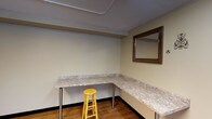 This single-floor space is designed with functionality and practicality at its core. The layout includes at least two distinct areas, each serving a specific purpose and finished with durable, low-maintenance materials.

The laundry room is equipped with a front-loading washer and dryer set, positioned side by side beneath bright fluorescent ceiling lighting. The room features warm beige walls, a window cutout for ventilation or pass-through access, and baseboard heating, making it a well-appointed utility area suited for regular use.

Adjacent to the laundry area is a dedicated work or vanity room that features an L-shaped countertop with a granite-style surface, supported by metal legs and offering generous workspace along two walls. Wood-look flooring adds warmth underfoot, while a framed wall mirror provides both a functional and decorative element. A simple stool completes the setup, making this area adaptable for a variety of uses, from grooming to crafting or administrative tasks.

Throughout the space, the walls are finished in a neutral beige tone, contributing to a clean and cohesive aesthetic. The overall design prioritizes utility and ease of use, with straightforward finishes and well-placed lighting ensuring each area is both functional and accessible.