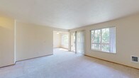 This single-floor residence offers a well-organized layout designed for comfortable everyday living. The living area is generously proportioned, featuring soft wall-to-wall carpeting and a neutral color palette that enhances the sense of openness. Multiple windows, including a large picture window and a sliding glass door with vertical blinds, flood the space with natural light and provide views of surrounding mature trees. The sliding door opens to an outdoor area, seamlessly connecting interior and exterior spaces. An adjacent dining area shares the same bright, airy atmosphere, accented by a ceiling fan fixture.

The kitchen is laid out in a practical U-shape, featuring wood-toned cabinetry with white cabinet faces, laminate countertops, and wood-look flooring. White appliances—including a refrigerator, electric range, dishwasher, and vent hood—provide a full suite of functionality.

The bathroom is finished with wood-look flooring and includes a combination tub and shower enclosed by sliding glass doors, a vanity with under-sink storage, and a large mirror. A separate bedroom offers plush carpeting and a built-in shelving unit with a desk surface, providing practical storage and workspace in a calm, neutral setting.