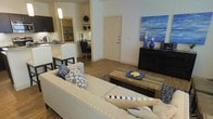 This single-floor residence features an open-concept layout that connects the living, dining, and kitchen areas into one cohesive and functional space. The living area is anchored by a generously sized cream sectional sofa accented with blue and white patterned throw pillows, complementing the space's consistent navy and neutral color theme. A rustic wood coffee table sits at the center atop a patterned area rug, adding textural contrast to the clean, modern aesthetic. Hardwood-style flooring runs throughout, contributing warmth and continuity across the space. Adjacent to the living area, a compact dining nook features dark bistro-style chairs positioned at a small table, tucked neatly beside the kitchen. The kitchen is outfitted with white cabinetry, light stone countertops, and a raised breakfast bar with dark bar stools, providing additional seating. Open shelving in the kitchen area offers display and storage space. A pendant light above the kitchen adds focused illumination. A dark dresser along the wall near the entryway serves as a decorative focal point, styled with blue accent pieces and wall art, reinforcing the space's unified, contemporary design.