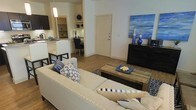 This single-floor residence features an open-concept layout that connects the living, dining, and kitchen areas into one cohesive and functional space. The living area is anchored by a generously sized cream sectional sofa accented with blue and white patterned throw pillows, complementing the space's consistent navy and neutral color theme. A rustic wood coffee table sits at the center atop a patterned area rug, adding textural contrast to the clean, modern aesthetic. Hardwood-style flooring runs throughout, contributing warmth and continuity across the space. Adjacent to the living area, a compact dining nook features dark bistro-style chairs positioned at a small table, tucked neatly beside the kitchen. The kitchen is outfitted with white cabinetry, light stone countertops, and a raised breakfast bar with dark bar stools, providing additional seating. Open shelving in the kitchen area offers display and storage space. A pendant light above the kitchen adds focused illumination. A dark dresser along the wall near the entryway serves as a decorative focal point, styled with blue accent pieces and wall art, reinforcing the space's unified, contemporary design.