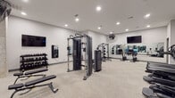 Located at 320 S Walker St in Cary, North Carolina, this fitness center offers a thoughtfully designed workout space that caters to a wide range of exercise needs. The room features neutral-toned walls and light carpet flooring, creating a clean and modern aesthetic throughout.

The strength training area is outfitted with TRUE-brand cable machines, a power rack with weight plates, a dumbbell rack with a full set of free weights, and flat workout benches. A large wall-mounted mirror spans one side of the room, allowing users to monitor their form during exercises. Wall-mounted flat-screen televisions provide entertainment throughout the space.

The cardio section includes multiple TRUE elliptical trainers with interactive digital displays, a treadmill, and a rowing machine, all arranged along the perimeter of the room to maximize open floor space. A Reflect interactive fitness mirror is mounted on the wall, offering guided workout programming as an added amenity.

Overhead recessed lighting illuminates the space evenly, while wall-mounted industrial fans provide air circulation during intense workouts. An exposed concrete column adds an architectural detail that contrasts with the otherwise sleek, contemporary interior. The overall layout is practical and spacious, offering ample room to move freely between equipment stations.