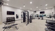 Located at 320 S Walker St in Cary, North Carolina, this fitness center offers a thoughtfully designed workout space that caters to a wide range of exercise needs. The room features neutral-toned walls and light carpet flooring, creating a clean and modern aesthetic throughout.

The strength training area is outfitted with TRUE-brand cable machines, a power rack with weight plates, a dumbbell rack with a full set of free weights, and flat workout benches. A large wall-mounted mirror spans one side of the room, allowing users to monitor their form during exercises. Wall-mounted flat-screen televisions provide entertainment throughout the space.

The cardio section includes multiple TRUE elliptical trainers with interactive digital displays, a treadmill, and a rowing machine, all arranged along the perimeter of the room to maximize open floor space. A Reflect interactive fitness mirror is mounted on the wall, offering guided workout programming as an added amenity.

Overhead recessed lighting illuminates the space evenly, while wall-mounted industrial fans provide air circulation during intense workouts. An exposed concrete column adds an architectural detail that contrasts with the otherwise sleek, contemporary interior. The overall layout is practical and spacious, offering ample room to move freely between equipment stations.