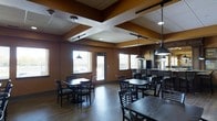 This single-floor dining space offers a comfortable and functional environment designed to accommodate groups of guests with ease. The room is finished in warm, earthy tones — deep amber and brown wall colors create an inviting ambiance that complements the dark wood furniture throughout. Sturdy dark-stained dining chairs and tables are arranged throughout the space, providing flexible seating configurations for various group sizes. Overhead, a combination of recessed lighting, track lighting, and industrial-style pendant lights hang at varying heights, casting a warm, even glow across the room. A distinctive coffered ceiling detail at the center of the space adds an architectural focal point, enhancing the overall design character. Natural light filters in through several windows fitted with horizontal blinds, offering views to the exterior while maintaining privacy. A service counter area is visible toward the rear of the dining room, suggesting a self-serve or staffed food and beverage station. The flooring throughout is a hardwood-style surface that adds warmth and durability. A glass-panel door provides additional access and contributes to the open, airy feel of the space. Overall, this dining area presents a welcoming and well-appointed setting suitable for everyday use or organized group dining.
