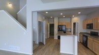 This three-story space is designed with a functional, open layout that promotes ease of movement and a cohesive flow between areas. The main level showcases an open-concept kitchen and living area, unified by warm hardwood-style flooring that extends throughout the space. The kitchen is equipped with light wood cabinetry, granite countertops, and a black refrigerator, offering both style and practicality. A kitchen island with a granite surface provides additional workspace and casual seating, making it a central hub for daily activities.

Recessed lighting is installed throughout the ceiling, ensuring consistent, well-distributed illumination across the main level. The neutral gray and white wall tones contribute to a clean, contemporary aesthetic that complements the natural wood tones of the flooring and cabinetry.

A staircase is visible near the entryway, leading to the upper floors, while a spacious walk-in storage area or pantry is accessible just off the main corridor. The architectural design features clearly defined structural columns and archways that add dimension without compromising the open feel of the space. Overall, the layout is practical, modern, and well-appointed for comfortable multi-level living.