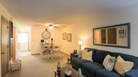 This two-floor residence offers a welcoming interior defined by warm tones and a practical open-concept layout on the main level. The living and dining areas flow seamlessly into one another, creating a cohesive shared space ideal for both relaxing and entertaining. Soft carpeting runs throughout, adding a cozy underfoot feel that complements the neutral, cream-toned walls. The living area is anchored by a navy blue sofa accented with decorative pillows, paired with a dark coffee table and accessorized with small décor pieces that add personality to the space. A large framed mirror on the wall enhances the sense of openness. The adjacent dining area accommodates a round table with white chairs, decorated with wall art and a large statement clock that adds a charming focal point. A ceiling fan with integrated lighting provides both air circulation and ambient illumination for the dining space, while a floor lamp in the corner contributes additional warm lighting to the living area. Natural light filters subtly through the space. The two-floor layout offers additional living or sleeping quarters on the upper level, providing a practical separation between common and private areas.