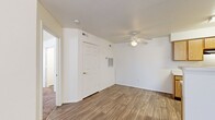 This single-story residential unit offers a practical and well-organized layout designed for comfortable everyday living. The heart of the home is an open-concept kitchen and living area, where warm-toned wood-look flooring extends throughout, creating a cohesive and inviting atmosphere. The kitchen features light oak cabinetry with ample storage, a white countertop peninsula with a built-in sink, and black appliances including a range and refrigerator, all beneath a textured ceiling. The living area is bright and open, benefiting from natural light streaming through a window fitted with white blinds, and includes a ceiling fan for added comfort. A dedicated dining or flex space adjacent to the kitchen also features a ceiling fan and continues the wood-look flooring. The bedrooms are carpeted for warmth and comfort, each offering generous proportions. The primary bedroom includes a wide sliding-door closet, a ceiling fan, and a window that allows natural light. Additional rooms provide versatile spaces suitable for various needs. The bathroom is clean and functional, featuring a full-size soaking tub with a shower surround. Throughout the unit, white six-panel doors, neutral white walls, and consistent finishes contribute to a clean, move-in-ready aesthetic.