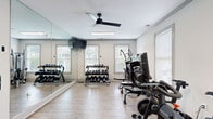 Located at 1001 Riverland Woods Pl in Charleston, SC, this fitness center offers a thoughtfully designed, single-floor workout facility that balances functionality with a clean, modern aesthetic. The space is organized into distinct activity zones, all unified by light wood-look flooring and crisp white walls that contribute to a bright, energizing atmosphere.

Upon entry, guests are welcomed by a striking living green wall featuring a motivational sign — a bold decorative statement that sets an encouraging tone. The cardio zone houses multiple treadmills and stationary cycling bikes, equipped with individual screens and positioned near windows that allow natural light to filter in. Ceiling fans overhead promote airflow throughout the space.

The strength training area features a comprehensive dumbbell rack alongside multi-use cable and weight machines, with floor-to-ceiling mirrors along one wall to maximize the sense of space and support proper form. A dedicated open-floor stretching and flexibility zone includes a massage chair, resistance bands, and yoga accessories, separated from the main floor by a sheer curtain partition for a more relaxed environment.

Throughout the facility, large windows with white blinds provide views of the surrounding greenery, reinforcing the connection to the natural Charleston landscape. The overall design is clean, well-lit, and purposefully equipped to support a full range of fitness activities.