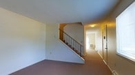 This two-story residence offers a functional and straightforward layout spread across two levels. The main floor features a carpeted living area finished in soft, neutral tones that create a calm and understated ambiance. A staircase with clean metal railings rises from the main level to the upper floor, serving as a central architectural element that divides the space while maintaining an open sightline. The textured ceiling adds a subtle layer of detail to the interior. A hallway extends from the base of the staircase, providing access to additional rooms at the rear of the main floor. Warm, ambient lighting illuminates the corridor, contributing to the overall welcoming feel of the space. A wall-mounted climate control unit is visible along the hallway, indicating the residence is equipped for year-round comfort. The use of consistent carpeting, light-colored walls, and modest finishes throughout gives the space a cohesive, practical aesthetic. The two-floor configuration provides clear separation between living and private areas, making this a well-organized and comfortable residential space suited for everyday living.