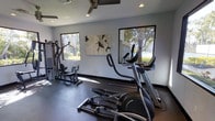 This single-floor fitness center offers a dedicated space for exercise and wellness, thoughtfully designed to accommodate a range of workout routines. The room features a dark rubberized flooring that provides both durability and comfort underfoot, ideal for high-impact activities and equipment use. Multiple large, square windows line the walls, flooding the space with natural light and offering pleasant views of the surrounding greenery and trees, creating an energizing atmosphere for exercise. A ceiling fan is centrally mounted overhead, ensuring consistent airflow throughout the room. The fitness area is equipped with a variety of cardio and strength training machines, including elliptical trainers and a cable/multi-use resistance machine, catering to a full-body workout experience. Recessed lighting supplements the natural light, keeping the space bright and well-lit at any time of day. A piece of abstract wall art adds a subtle decorative touch, balancing the functional nature of the room with a sense of style. The overall ambiance is clean, modern, and practical, making it a welcoming environment for fitness enthusiasts of all levels.