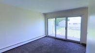 This single-floor space features a bright and simply designed living area that emphasizes functionality and comfort. The room is finished with wall-to-wall carpeting in a neutral tone, providing a soft, comfortable underfoot feel throughout. The walls are painted in a clean, light cream hue, contributing to an open and airy atmosphere.

A standout feature of this space is the large sliding glass door unit that spans a generous portion of one wall, flooding the interior with natural light and providing direct access to a private balcony. The balcony is enclosed by a white railing and overlooks a lush, green outdoor setting, offering pleasant views of surrounding greenery and neighboring structures.

Baseboard heating runs along the base of the exterior wall, providing an efficient heating solution well-suited for cooler climates. The overall layout is open and uncluttered, making it highly adaptable for a variety of furniture arrangements. The combination of natural light, outdoor access, and neutral finishes creates a welcoming, versatile living environment.